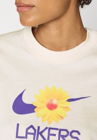 Grietinėlių spalvos sportinis džemperis su geltonu gėlių ir krepšinio grafiku virš violetinio Nike logotipo, o apačioje - žodis "LAKERS" piksliškoje tekstūroje.