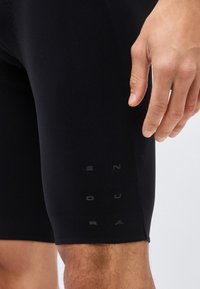 Κοντινό του χεριού ενός ατόμου που ακουμπά δίπλα σε μαύρα εφαρμοστά σορτς με διακριτικό κάθετο κείμενο στον μηρό.