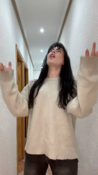 Kvinde med langt sort hår iført en beige sweater løfter begge hænder, mens hun kigger opad i en smal, veloplyst gang med trædørklinker.