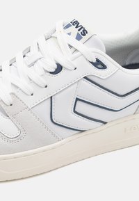 Vit sneaker tillverkad av läder och mocka, med en grå mockapanel, blå accenter, runda skosnören och en texturerad gummisula.