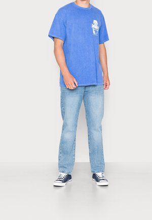 Blaues Baumwoll-T-Shirt mit Taschen-Design und weißem Druck, kombiniert mit hellblauen Jeans und navyfarbigen Leinenschuhen.