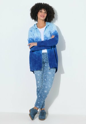 Vrouw met krullend haar die een blauwe tie-dye hoodie, wit shirt, blauw gebloemde broek en blauwe sneakers draagt, staand met gekruiste armen.