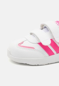 Zapatilla blanca con acentos rosas y diseño de tres franjas. Cuenta con correas de Velcro y una superficie texturizada para mayor agarre. Forma de punta redondeada.