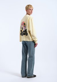 Langärmliges, hellgelbes Baumwollhemd mit floralem Grafikdesign und schwarzem Text auf dem Rücken. Kombiniert mit hellblauen Jeans.