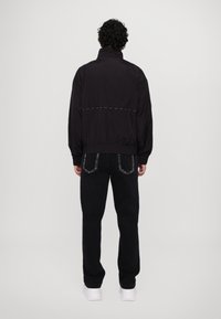 Schwarze leichte Jacke mit hohem Kragen, elastischen Bündchen und einem kontrastierenden, genähten Detail über den Rücken, kombiniert mit schwarzen Jeans.