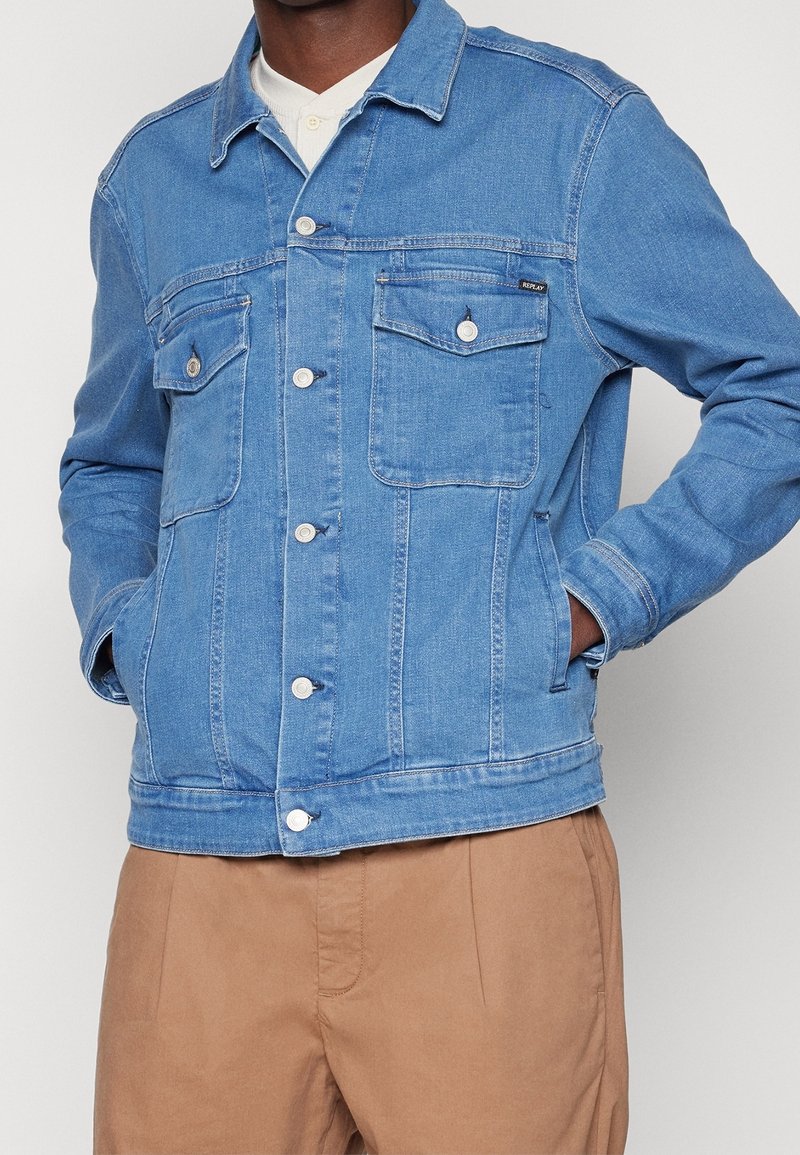 Man wearing a light blue denim jacket with buttoned chest pockets and hands in side pockets, paired with beige pants.