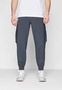 Pantaloni cargo da uomo grigio scuro con polsini elasticizzati, dotati di tasche laterali e un design elegante e leggero. Indossati con una maglietta bianca e sneakers.