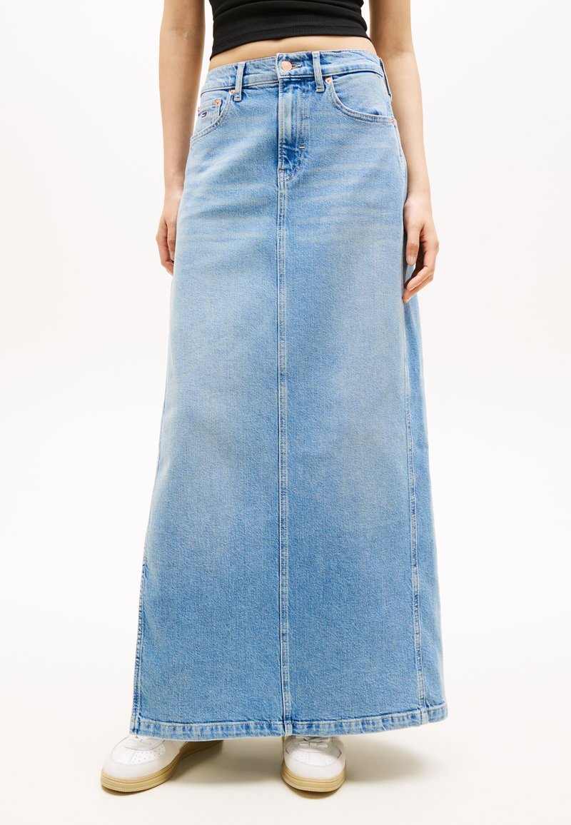 Άτομο που φοράει μακριά φούστα από ανοιχτό μπλε τζιν, μαύρο κομμένο τοπ και άσπρα αθλητικά παπούτσια, στέκεται μπροστά σε ένα απλό λευκό φόντο.