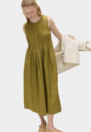 Donna che indossa un abito midi verde oliva senza maniche con pieghe, mentre si mette o si toglie una giacca beige chiaro davanti a uno sfondo semplice.