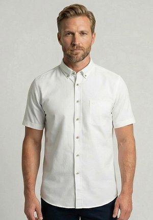 Blonde man met baard die een wit overhemd met korte mouwen, knopen en borstzak draagt, staande tegen een effen lichte achtergrond.