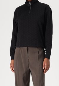 Pullover noir court avec un col montant et une fermeture éclair quart. Présente un motif texturé et des poignets ajustés, associé à un pantalon gris.