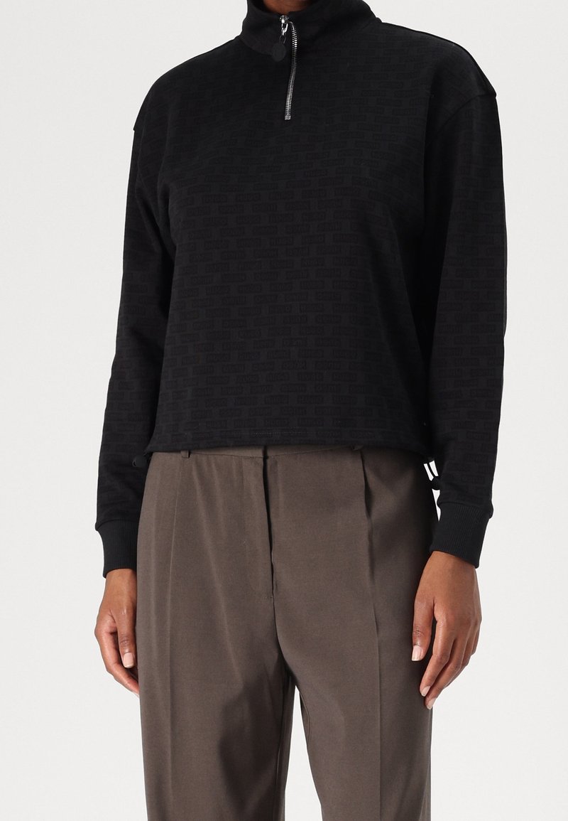 Pullover noir court avec un col montant et une fermeture éclair quart. Présente un motif texturé et des poignets ajustés, associé à un pantalon gris.