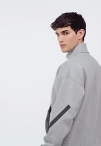 Grå sweatshirt med hög krage, avslappnad passform, långa ärmar och en svart accent på vänster ärm. Slät textur.