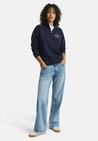 Marineblauwe, lange mouwen quarter-zip pullover in katoenmix, met witte geborduurde tekst. Gecombineerd met lichtblauwe wijde jeans en beige sneakers.