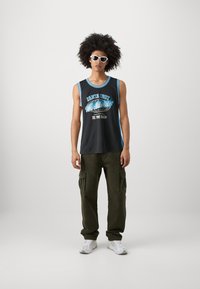 Jersey negro sin mangas con ribete azul, que presenta el texto "Santa Cruz Worldwide" en impresión estilizada. Combinado con pantalones cargo oliva y zapatillas blancas.
