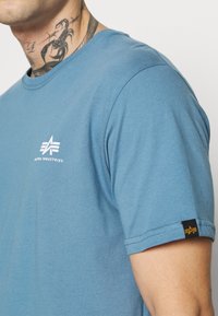 Kurzärmliges blaues T-Shirt aus weichem Stoff, mit einem weißen Logo auf der Brust und einem schwarzen Etikett mit gelben Akzenten am Ärmel.