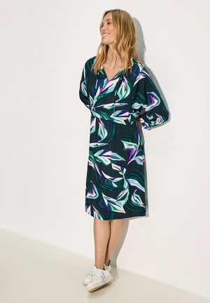 Kleid mit floralem Druck und tiefem V-Ausschnitt, gepufften Ärmeln, lebhaftem Teal, Lila und Weiß auf dunklem Hintergrund; kombiniert mit weißen Sneakers.