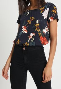 Blusa con estampado floral que presenta mangas cortas, un fondo azul marino y flores vibrantes en rojo, blanco y verde, combinada con jeans negros de cintura alta.