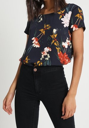 Blouse à motif floral avec des manches courtes, un fond marine, et des fleurs rouges, blanches et vertes éclatantes, associée à un jean noir taille haute.