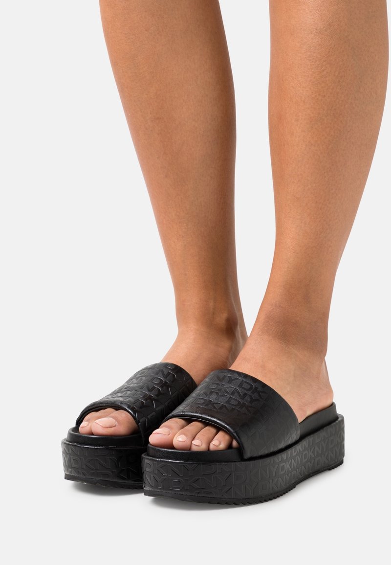 Pieds portant des sandales plateformes noires à enfiler avec des brides texturées et des semelles embossées du logo DKNY sur un fond clair.