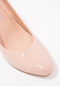 Pompe en cuir verni rose brillant avec un bout rond et un design minimaliste. L'intérieur présente une doublure beige lisse et un léger talon.