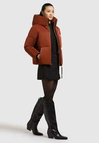 Giacca puffer di un arancione ruggine con colletto alto, design trapuntato e chiusura con zip, abbinata a una minigonna nera e stivali neri al ginocchio.