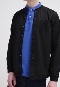Chaqueta negra de botones sobre una camiseta polo azul, con cuello redondo y un solo bolsillo en el pecho. El material parece suave y tiene un ajuste casual.