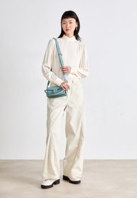 Creme højhalset bluse, brede beige bukser og kraftige sorte såler i hvide sko. Modellen bærer en lille, struktureret lyseblå crossbody-taske.