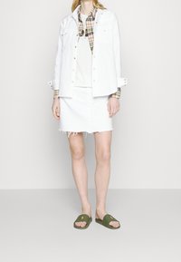 En person som bär en vit skjorta med knappar över en rutigt skjorta och en t-shirt med tryck, vit kjol med fransad fåll och gröna slidesandaler med orange logotyper.