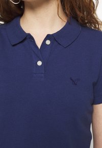 Námornícky polokošile vyrobená z bavlny, obsahující límec, zapínání na dva knoflíky a erb orla v tmavě modré barvě na levé hrudi.
