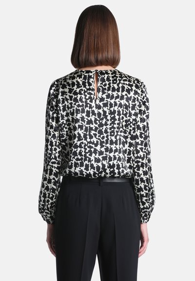 Femme aux cheveux bruns raides portant un chemisier à manches longues noir et blanc à motifs et un pantalon noir, vue de dos sur fond blanc.
