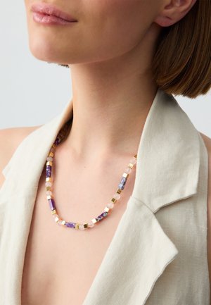 Collier en perles composé de composants violets, roses et dorés avec des formes rectangulaires et carrées. Porté sur un vêtement en lin de couleur claire.