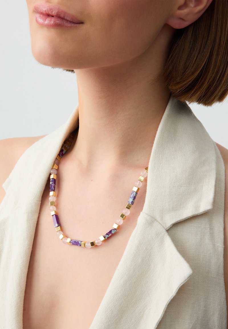 Collana di perline con componenti viola, rosa e oro, caratterizzata da forme rettangolari e quadrate. Indossata su un indumento in lino di colore chiaro.