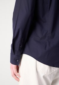 Personne portant une chemise à manches longues bleu marine foncé rentrée dans un pantalon blanc, montrant un gros plan du bras gauche, du poignet et de la main.