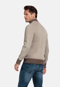 Hellbrauner Strickpullover mit dunkelbraunen Akzenten am Kragen und an den Bündchen. Hat ein figurbetontes Design, gezeigt von hinten, kombiniert mit blauen Jeans.