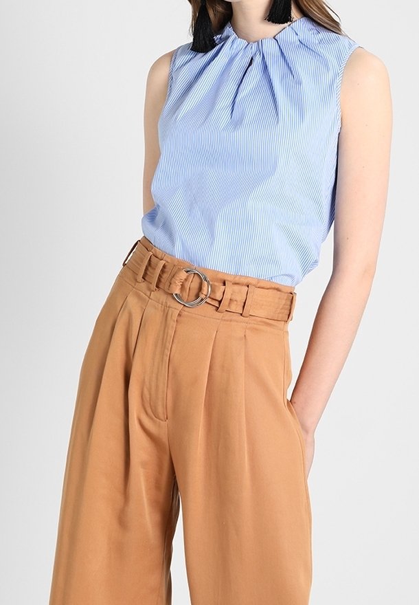 Femme portant un chemisier sans manches rayé bleu clair, rentré dans un pantalon large taille haute beige avec une ceinture à boucle ronde en argent.