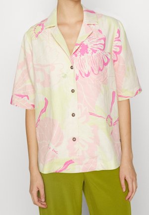 Femme portant une chemise à manches courtes boutonnée avec un motif floral rose pastel, jaune et blanc, et un pantalon vert olive.
