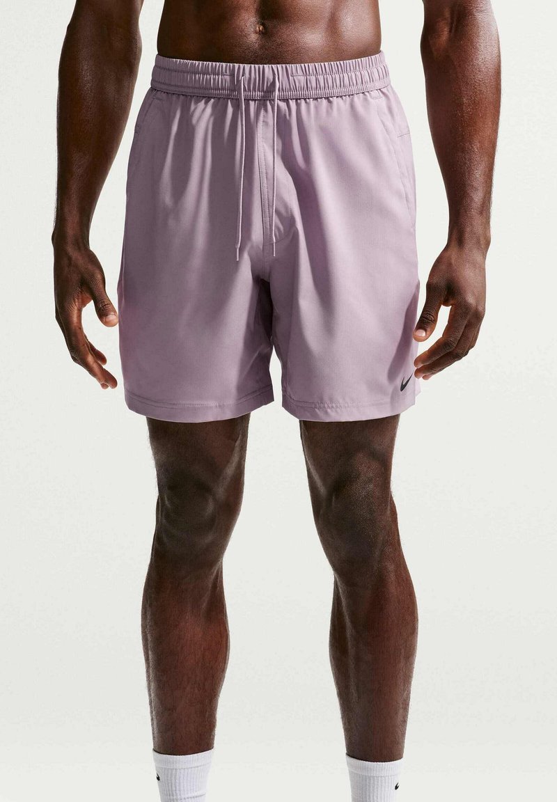 Homme athlétique portant un short Nike violet clair avec cordon et des chaussettes blanches Nike, debout devant un fond blanc uni.