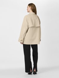 Beige trenchcoat med høy krage og bakflap, kombinert med svarte bukser med sideåpninger, båret med svarte hæler.