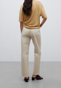 Kvinna med långt mörkt hår, iklädd beige byxor, en lös ljusbrun skjorta och svarta klacksandaler, står mot en enkel ljus bakgrund.