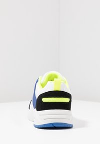 Sneaker blu e bianco con un accento nero testurizzato, una linguetta del tallone gialla brillante e un collare imbottito. Presenta una suola decorata per una migliore aderenza.