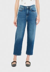 Jeans azules de talle alto y pernera recta con costuras en contraste, cinco bolsillos y cierre de botón. Combinados con bailarinas negras.