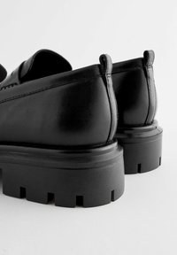 Scarpe in pelle nera con suola a piattaforma spessa, caratterizzate da una texture liscia e un tacco arrotondato, cuciture visibili e linguette sul retro.