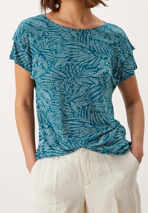 Frau trägt ein türkis gemustertes T-Shirt mit kurzen Ärmeln, das in beige Leinenhose mit hohem Bund gesteckt ist, die Hände in den Taschen.