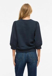 Femme aux longs cheveux blonds portant un haut bleu marine à manches bouffantes et un jean en denim bleu foncé, debout de dos face à la caméra.