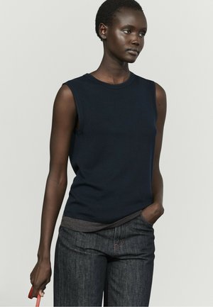 Donna con i capelli corti che indossa una canotta blu navy e jeans scuri, tiene una cinghia rossa in una mano e l'altra mano in tasca.