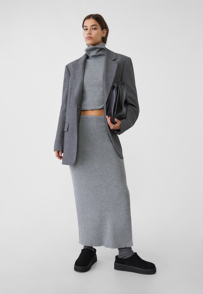 Graues, geripptes Strick-Zweiteiler-Set mit einem kurz geschnittenen Langarm-Oberteil und einem bodenlangen, eng anliegenden Rock, kombiniert mit einem grauen Blazer und schwarzen Schuhen.