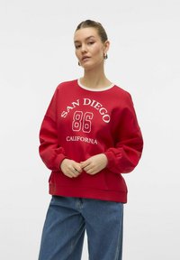 Sweat-shirt rouge avec le texte blanc "SAN DIEGO 86 CALIFORNIA", col rond, poignets côtelés, et coupe décontractée. Associé à un jean bleu.