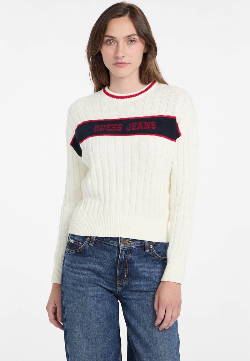 Pulover alb de tricot ribbit, cu o bandă orizontală largă bleumarin pe care este scris "GUESS JEANS" cu litere roșii. Este acompaniat de blugi închiși la culoare.