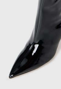Stivale alla caviglia in pelle verniciata nera con punta affusolata, caratterizzato da una finitura lucida e una texture liscia, evidenziato da dettagli di cucitura sottili.